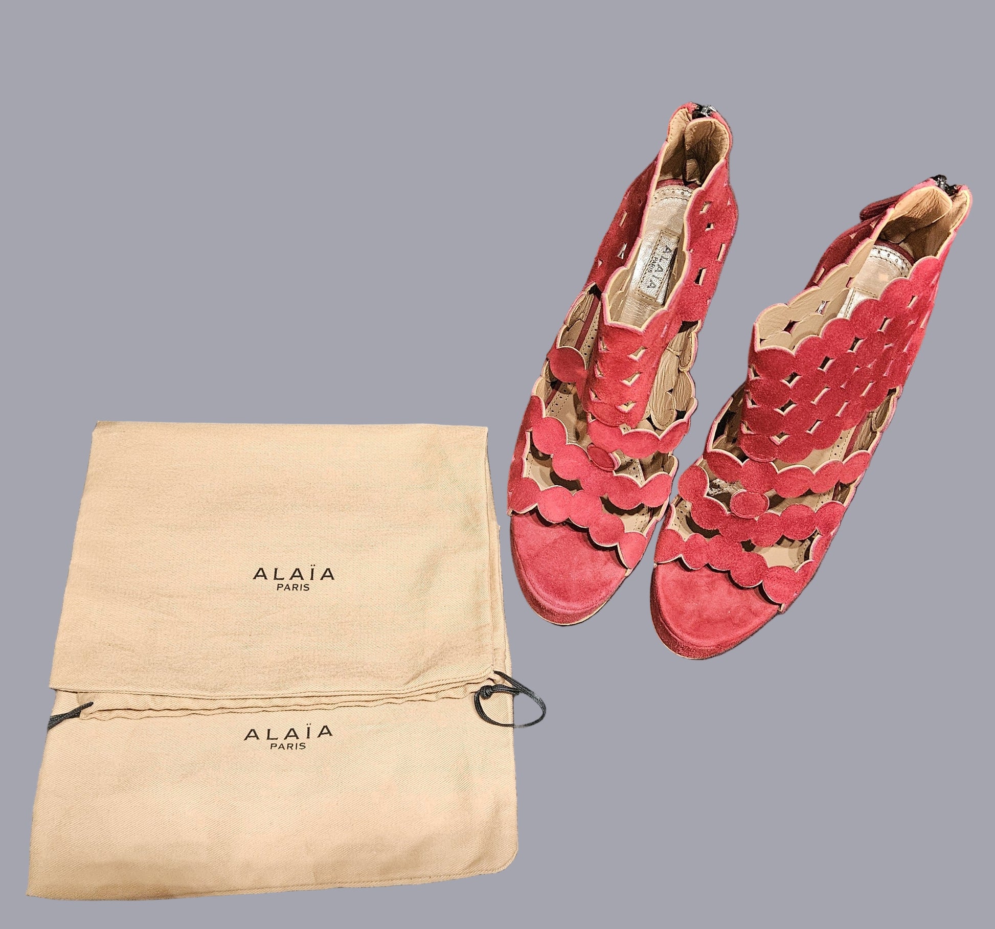 Red suede shoes with cut-out design next to an Alaia Paris dust bag on a gray surface.
