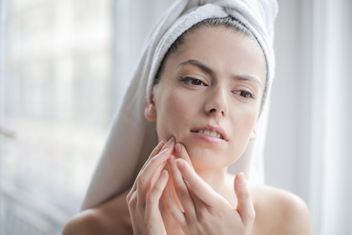 A woman with a white towel wrapped around her head, checking blemishes on her face