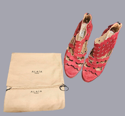 Red suede shoes with cut-out design next to an Alaia Paris dust bag on a gray surface.